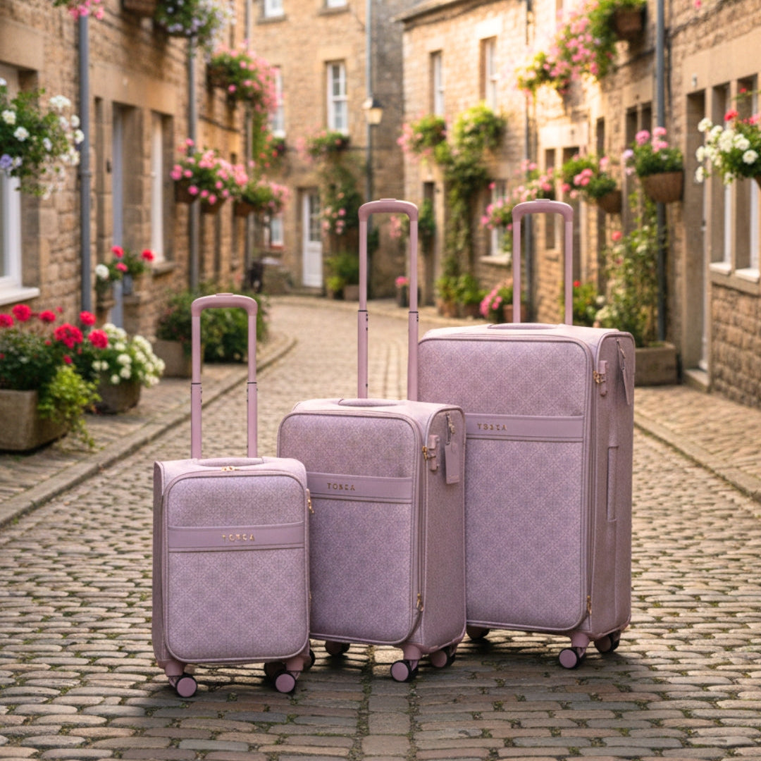 Set of three misty rose willow suitcases on a cobblestone street with floral decorations.